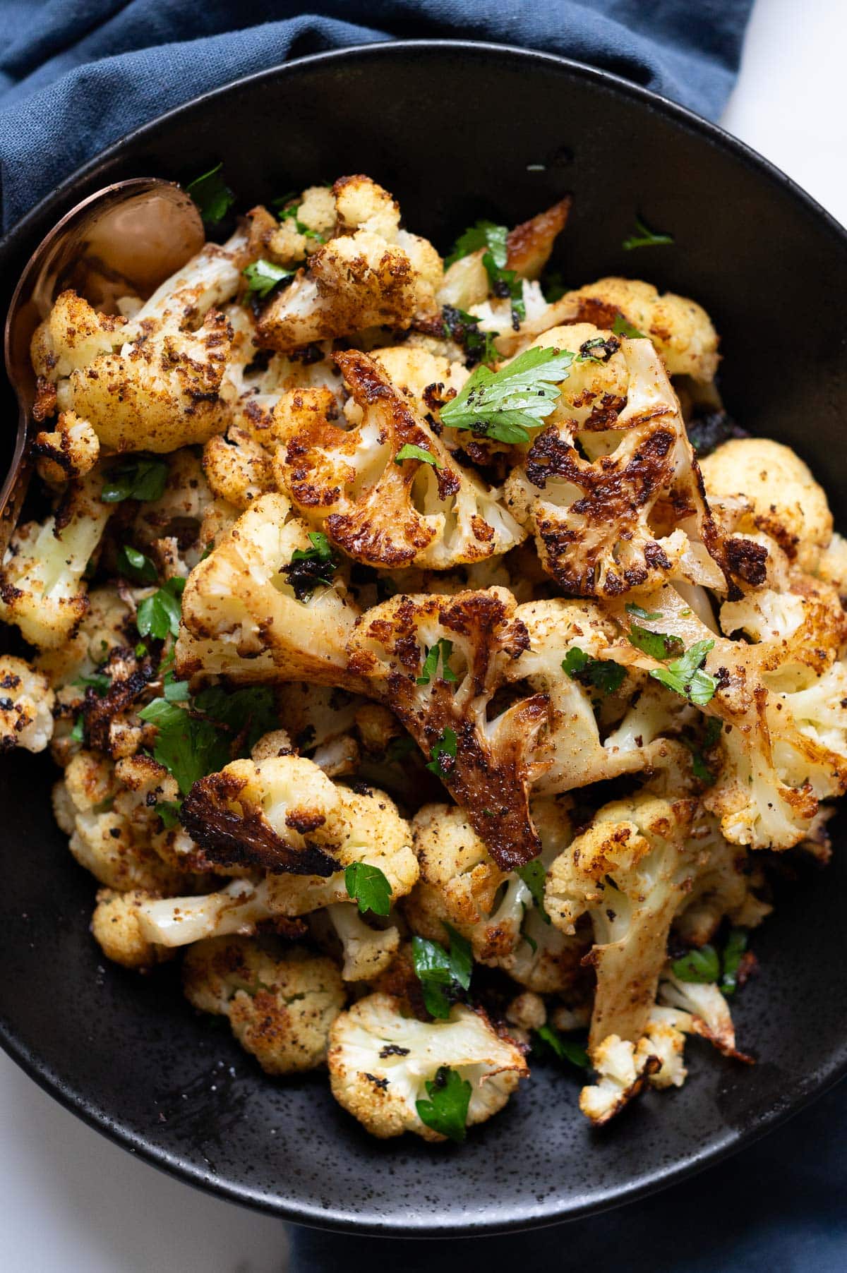 Roasted cauliflower in a bowl with a spoon. Blue linen napkin on a counter.