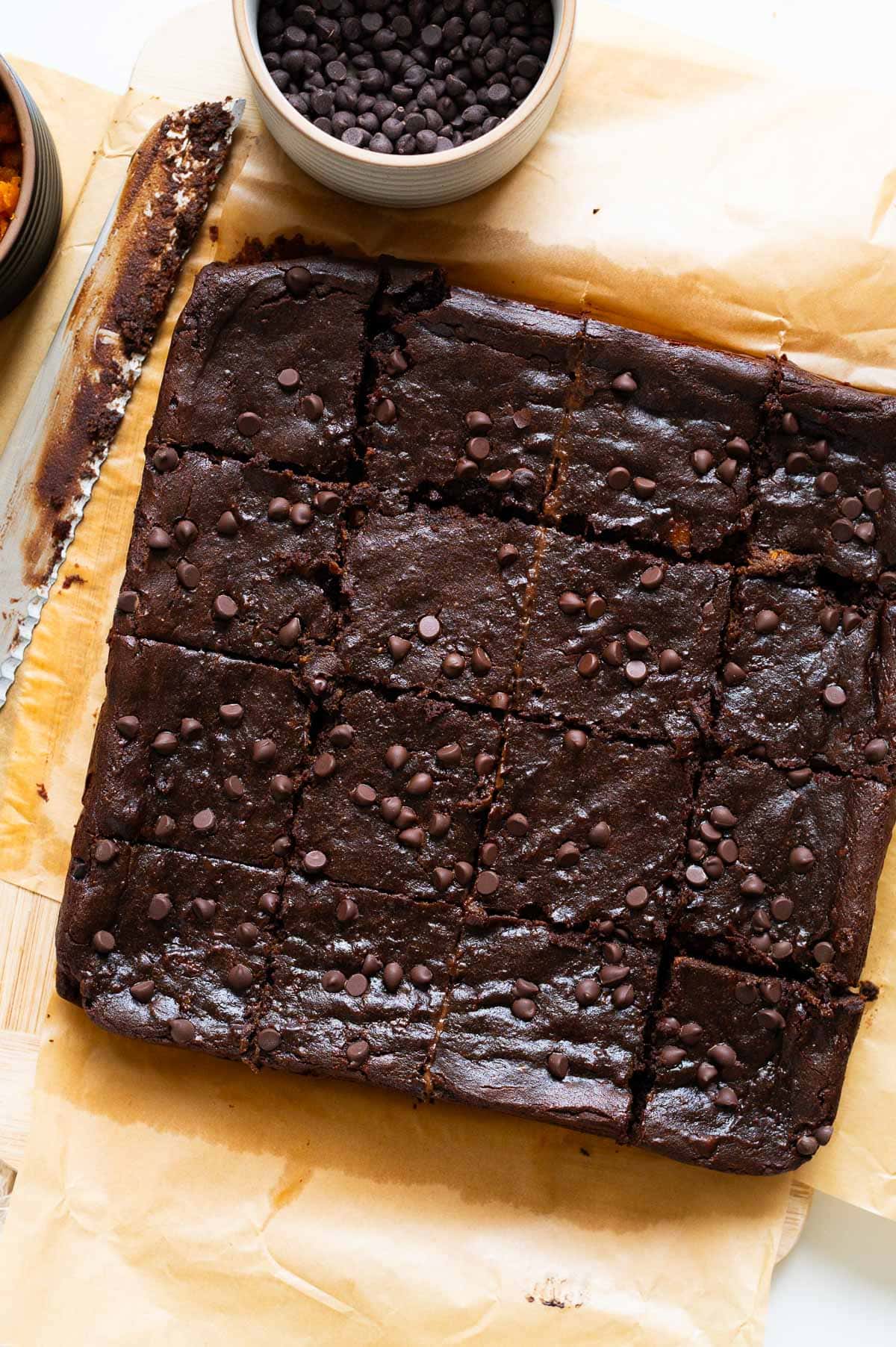 Sliced brownies on a parchment paper. Knife and bowl with chocolate chips next to it.