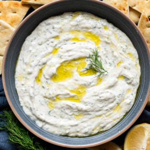 Tzatziki sauce drizzled with olive oil and garnished with dill in a bowl. Pita bread around it.