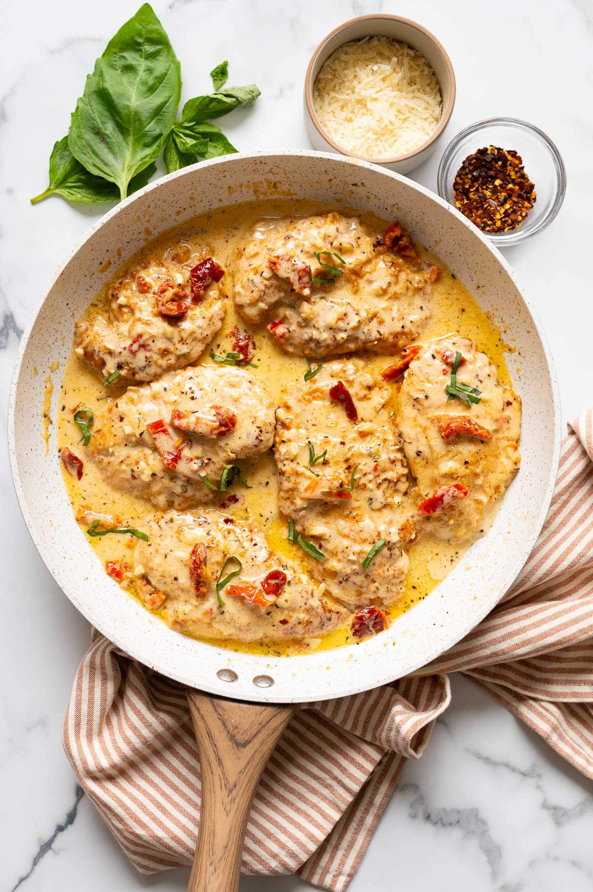 Marry me chicken in a skillet. Kitchen towel, basil, red pepper flakes and Parmesan cheese on a counter.