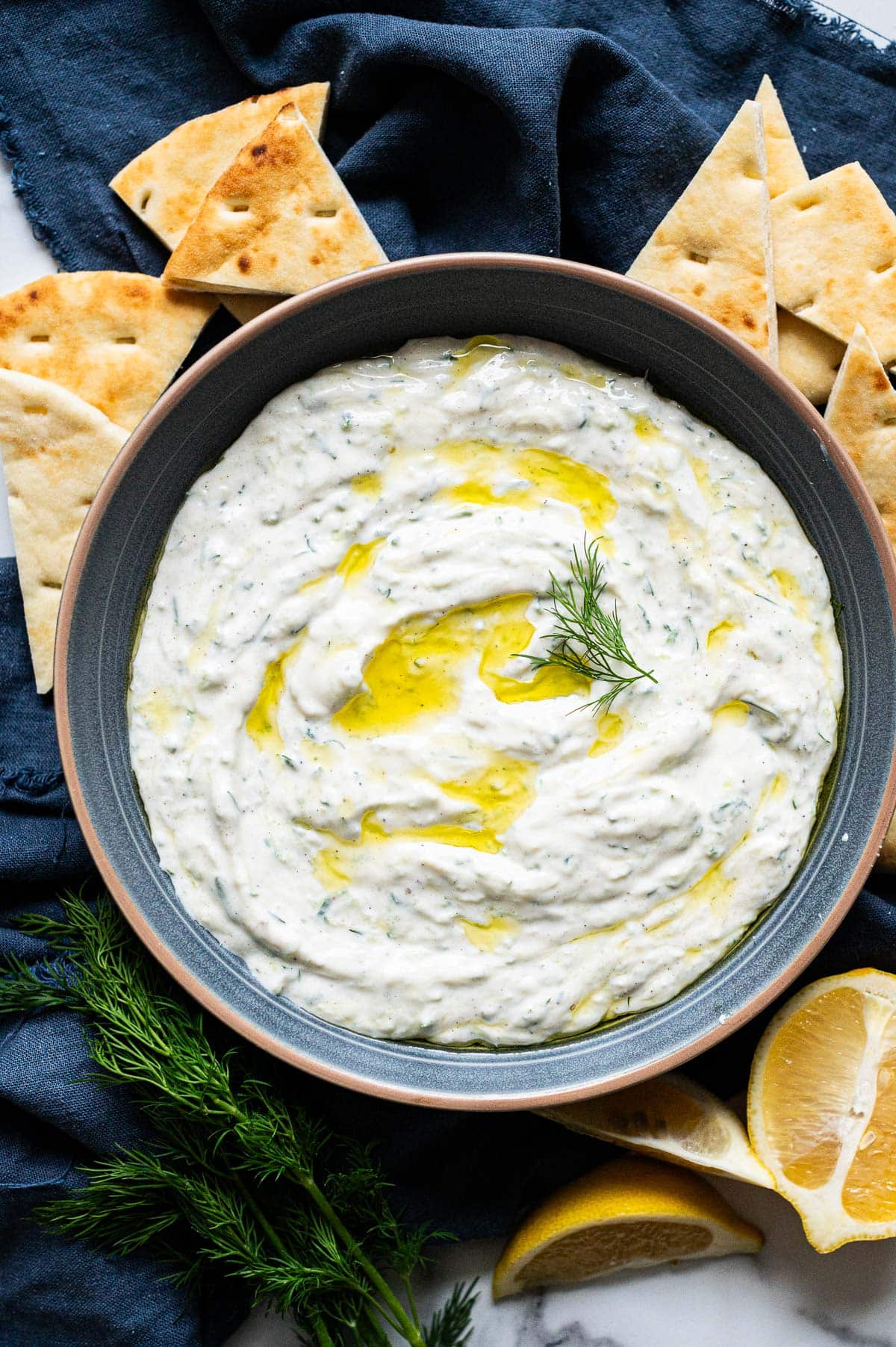 Tzatziki sauce drizzled with olive oil and garnished with dill in a bowl. Pita bread around it.