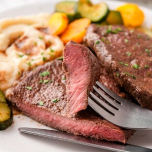 A slice of sirloin tip steak on a fork served with mashed potatoes and vegetables.