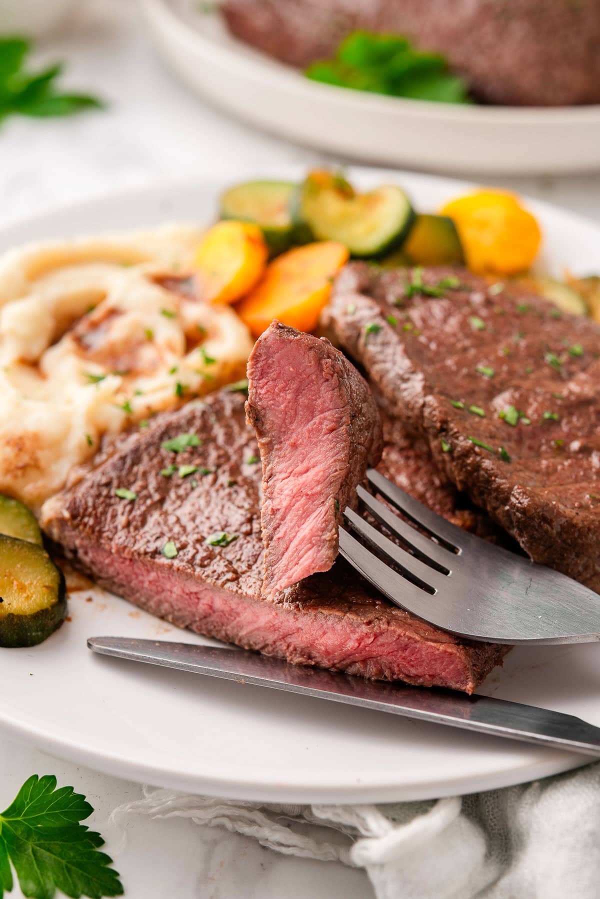 A slice of sirloin tip steak on a fork served with mashed potatoes and vegetables.