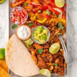 Sheet pan chicken fajitas with tortillas, pico de gallo, lime, sour cream and avocado.