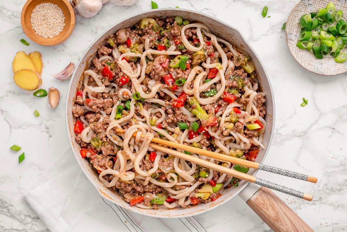 Ground beef stir fry with udon noodles in a skillet with chopsticks.