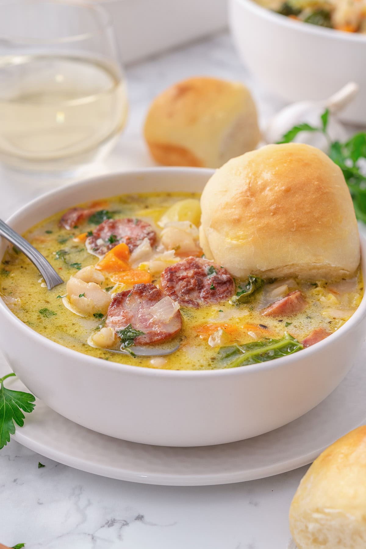 Sausage kale white bean soup in a bowl with a spoon and a bun.