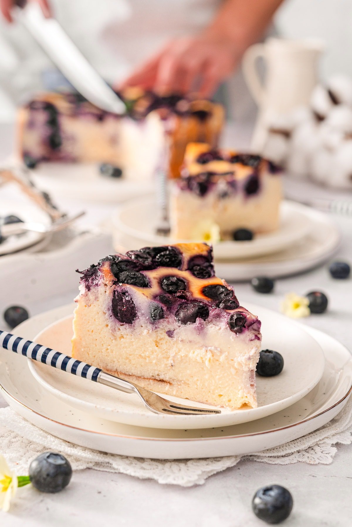 A slice of blueberry yogurt cake on a plate. Another slice and person cutting the cake behind.